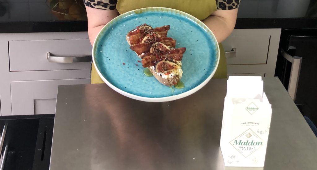 Woman holding plate of bacon and ricotta tartine with chilli with box of Maldon sea salt