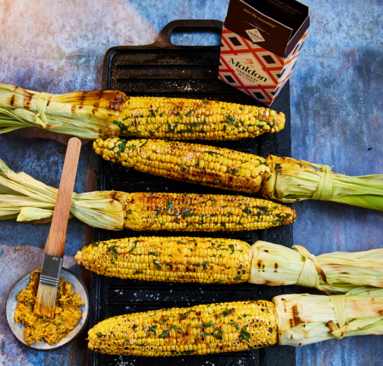 Four pieces of BBQ sweetcorn that have been flavoured with harissa and coriander butter on a griddle pan next to a box of Maldon Salt