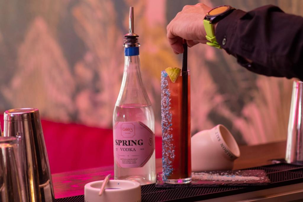 A sea breeze cocktail is stirred with a straw, placed next to a bottle of Spring Vodka and a salt pig.
