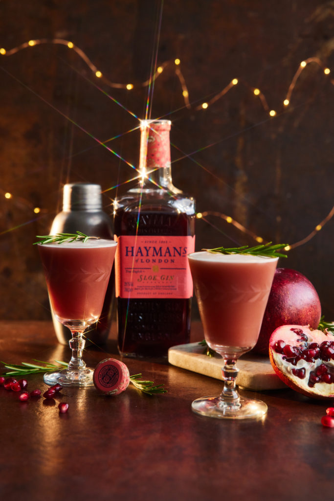 Two cocktails in front of a bottle of Hayman's sloe gin, Maldon Salt, and a cut pomegranate.