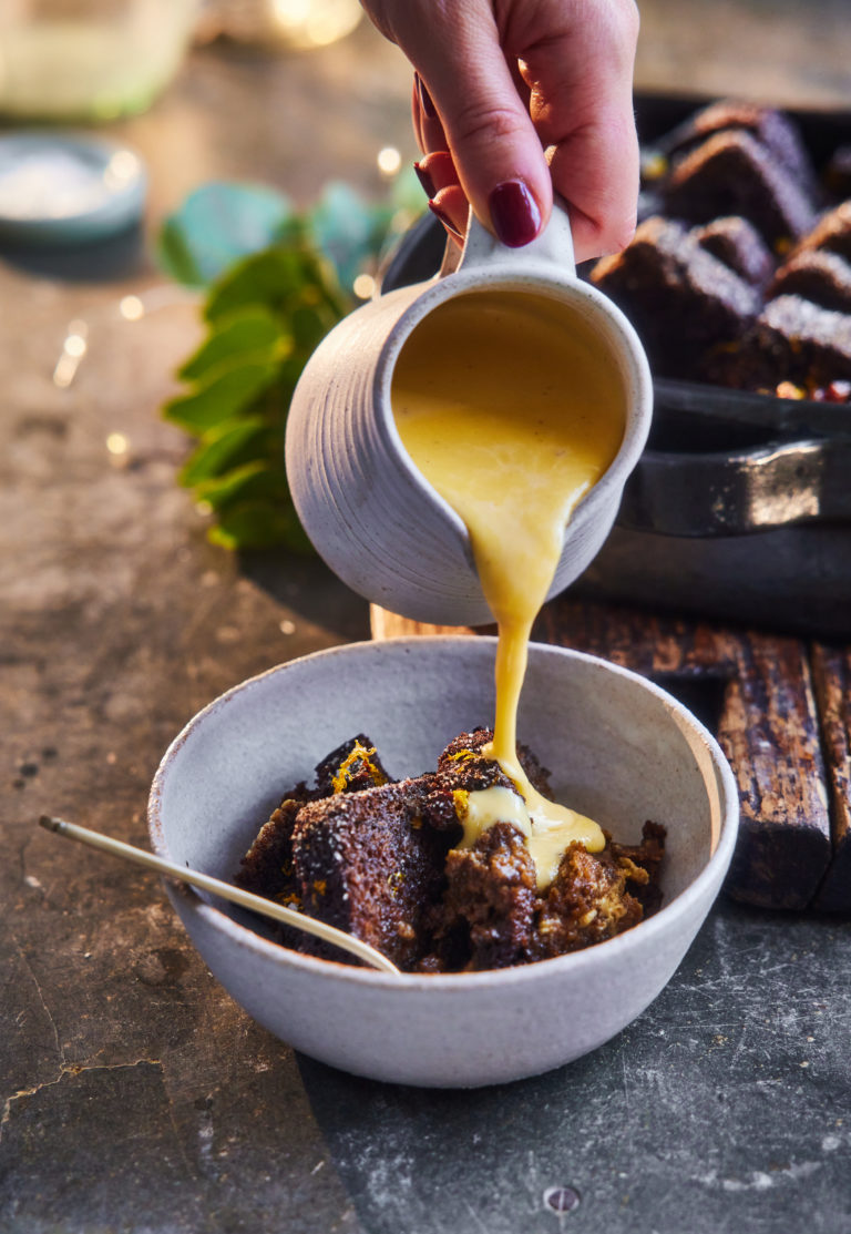 Gingerbread and butter pudding in a deep dish, next to a bowl of pudding topped with custard and a jar of custard.