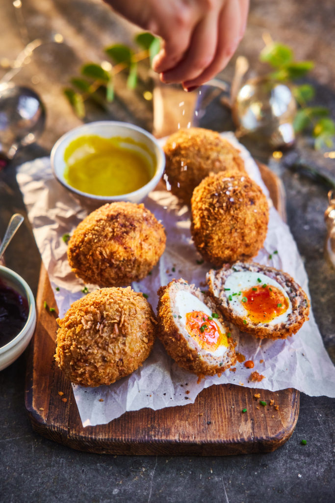Several turkey scotch eggs, with one cut open. A hand sprinkles salt over the eggs.