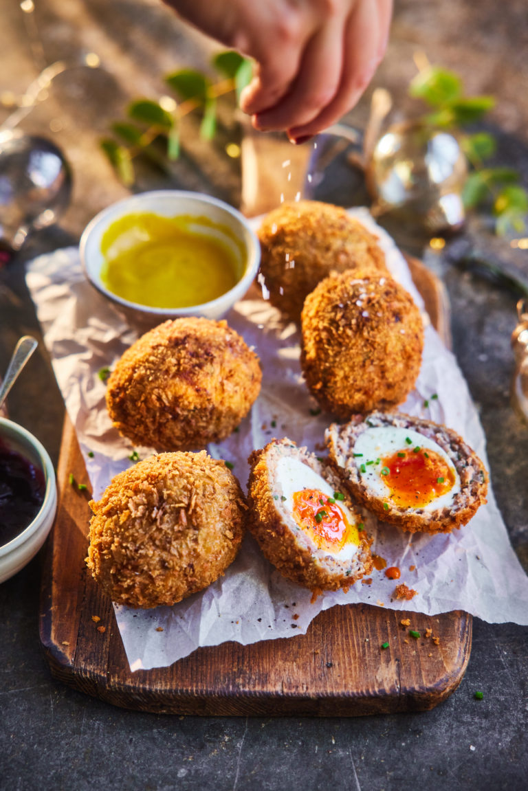 Several turkey scotch eggs, with one cut open. A hand sprinkles salt over the eggs.
