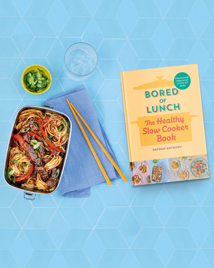 Metal mess tin containing noodles and peppers sits next to a small bow of spring onions, chopsticks, a glass of water and a copy of Bored of Lunch: The Healthy Slow Cooker Book.