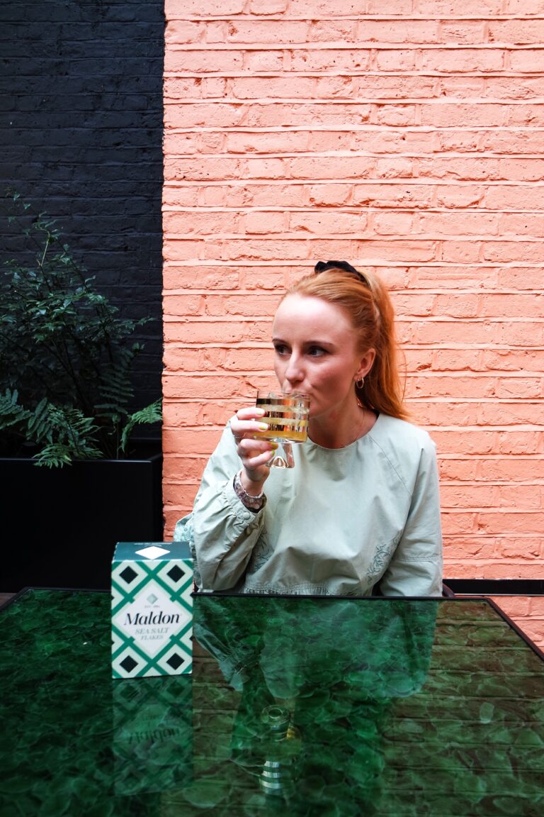 Imogen sits at a green table taking a sip from a glass.
