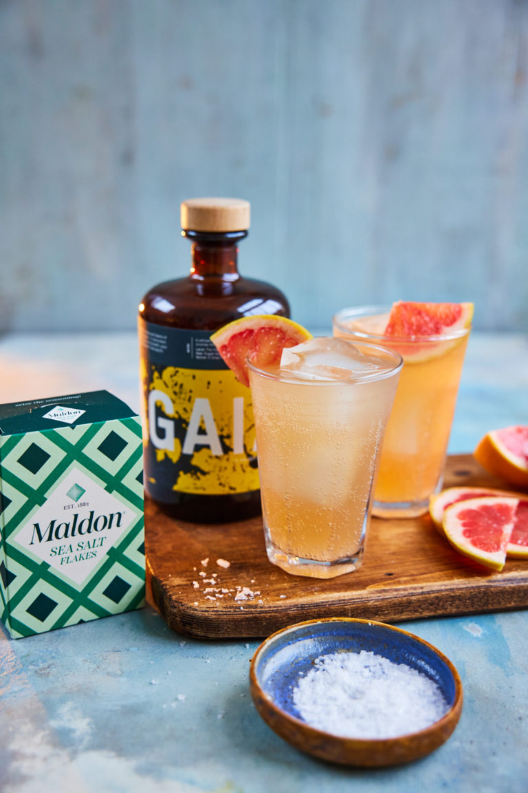 Two cocktails on a wooden board, next to sliced grapefruit, a box of Maldon Salt and a bottle of Feragaia.
