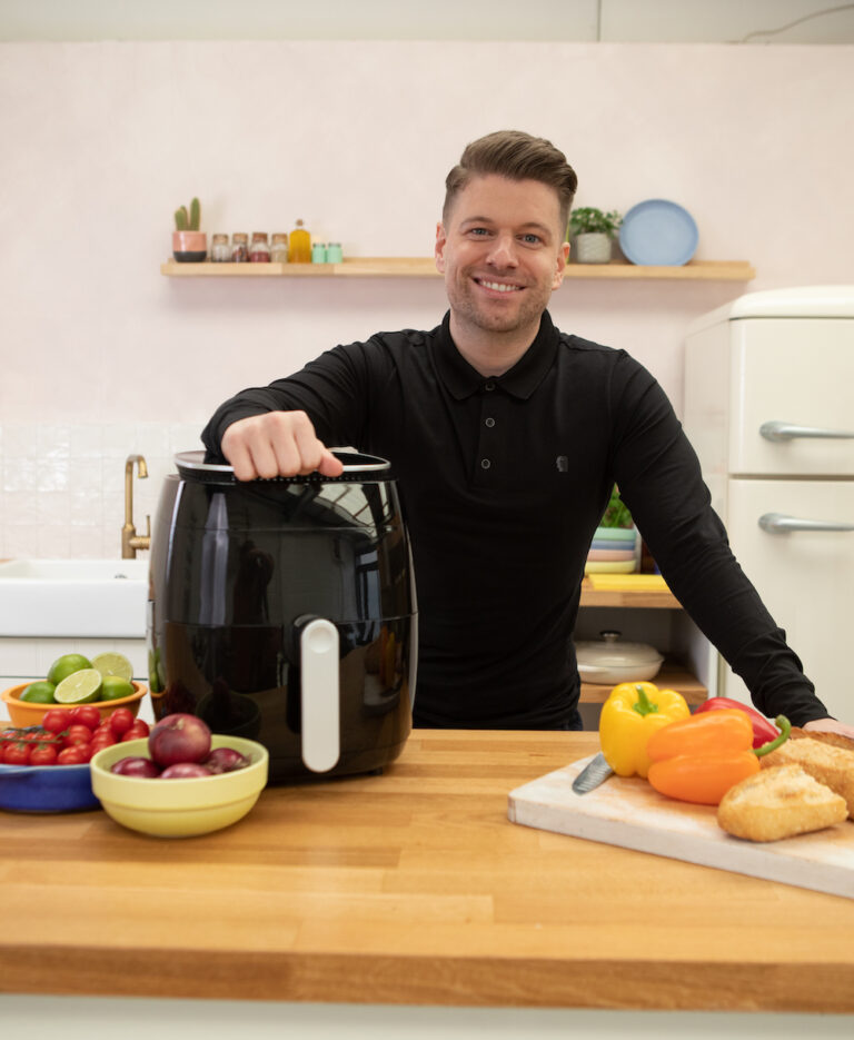 Nathan poses smiling behind a wooden counter with his arm resting on an air fryer.