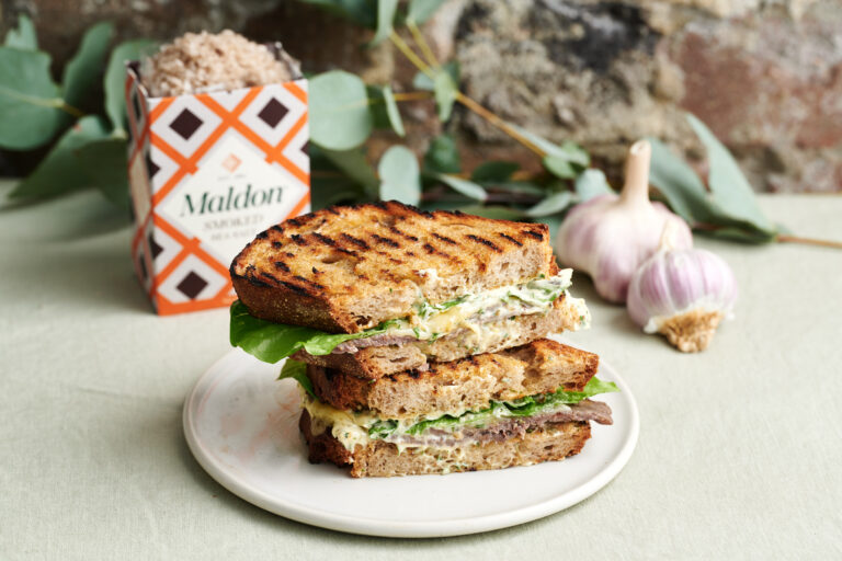 A steak sandwich cut in half, stacked up on a plate on a dinner table with Maldon Smoked Salt.
