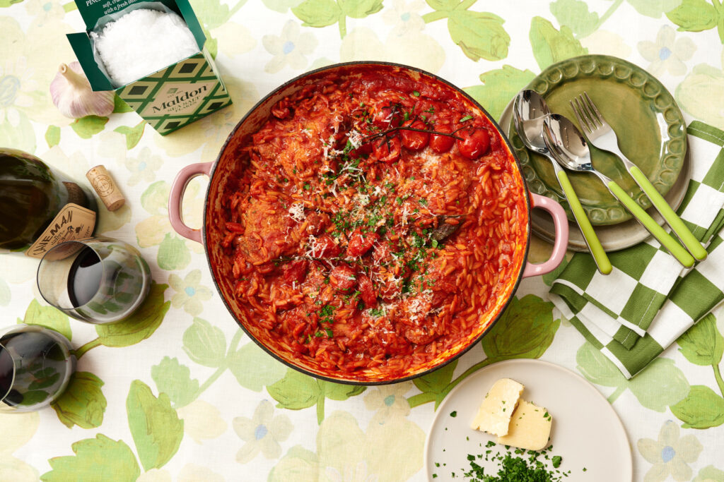 A pan of chicken risoni, photographer overhead, sitting upon a green floral tablecloth with 2 glasses of red wine.
