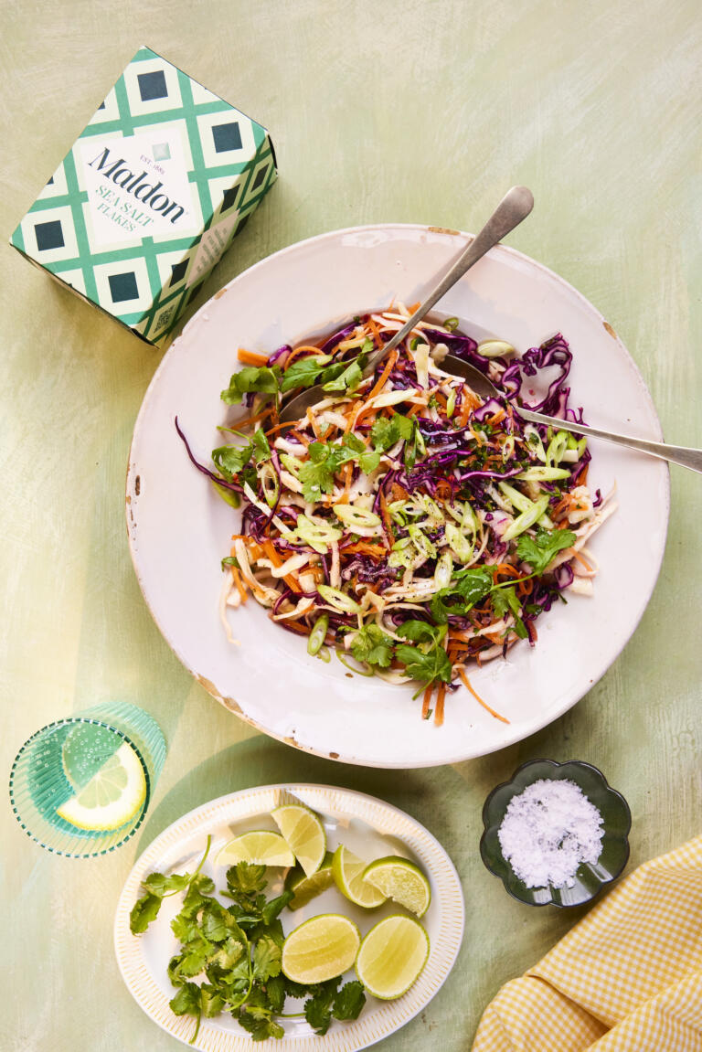 A bright, colourful cabbage coleslaw mixed with limes and coriander. Colours of red, orange, green and purple can be seen from the cabbage. Lime wedges and a dish of Maldon Sea Salt sit beside the dish.