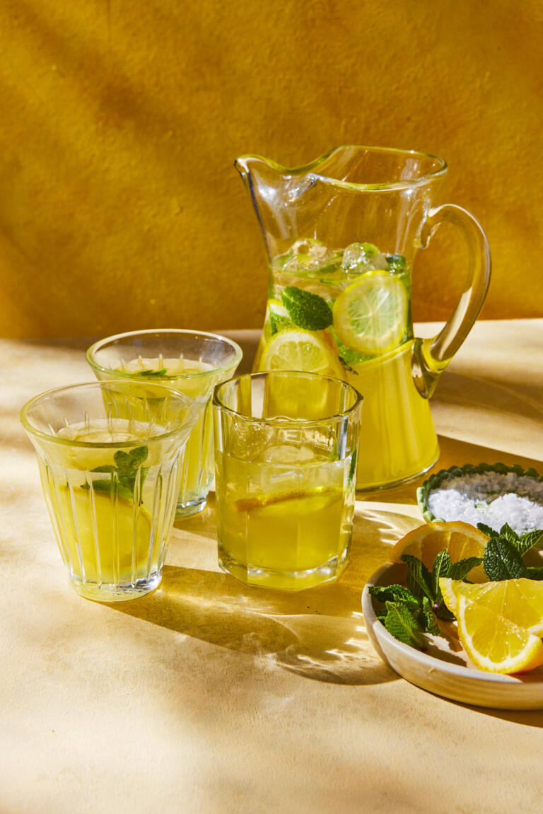 A large jug of homemade, golden lemonade with lemon slices and mint leaves floating in it. Three drinking glasses beside the jug which are filled to the top with the homemade lemonade. A dish of Maldon Sea Salt can be seen as well as a few lemon wedges.
