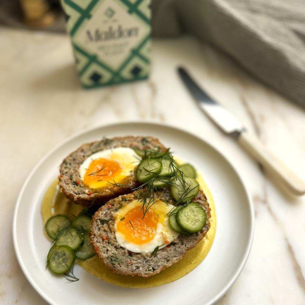 Sliced Scotch egg with a soft yolk, served with cucumber slices and dill on a white plate.